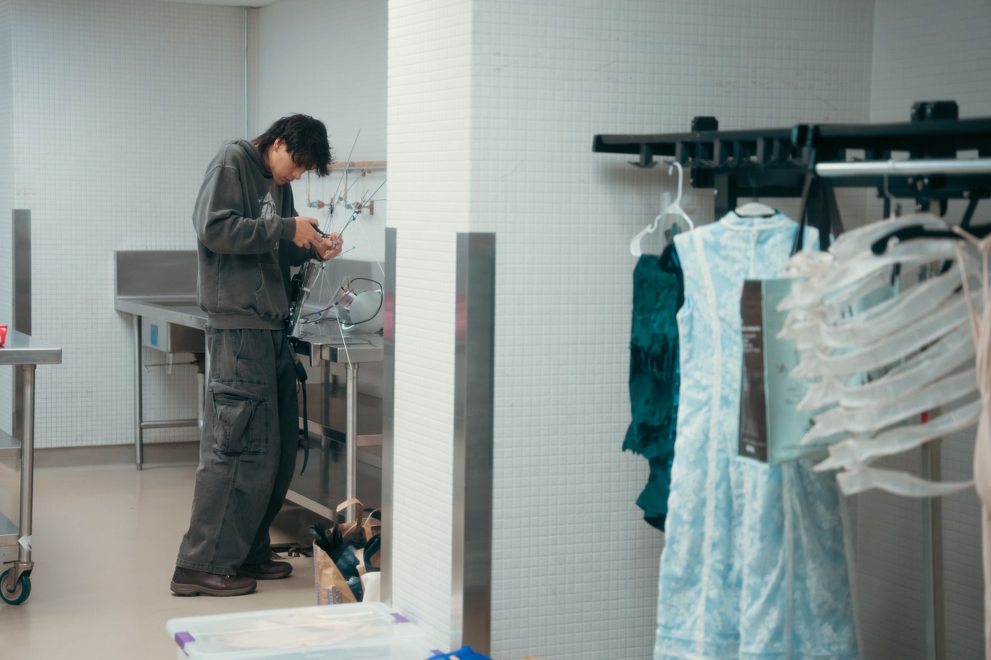Designer works on assembling a mechanical wearable in a backstage or workshop setting. They adjust metal rods connected to a harnessed structure with a spherical component on the table. Nearby, a rack displays garments, including a translucent sculptural dress.